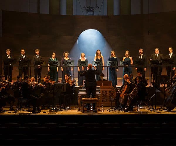 a group of 12 singers stand behind an orchestra with an illuminate circle behind