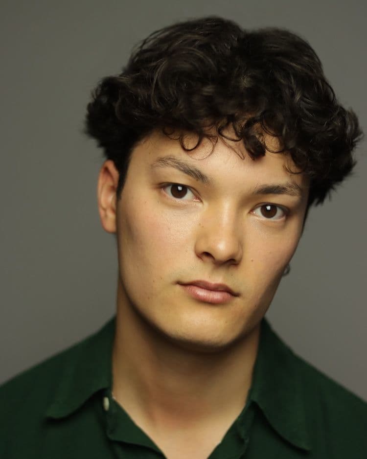 headshot of Actor Ty Arnott, looks at camera, a young man with curly dark brown hair and brown eyes. He has a neutral expression, is wearing a dark green collared shirt, and is posed against a solid, neutral gray background.