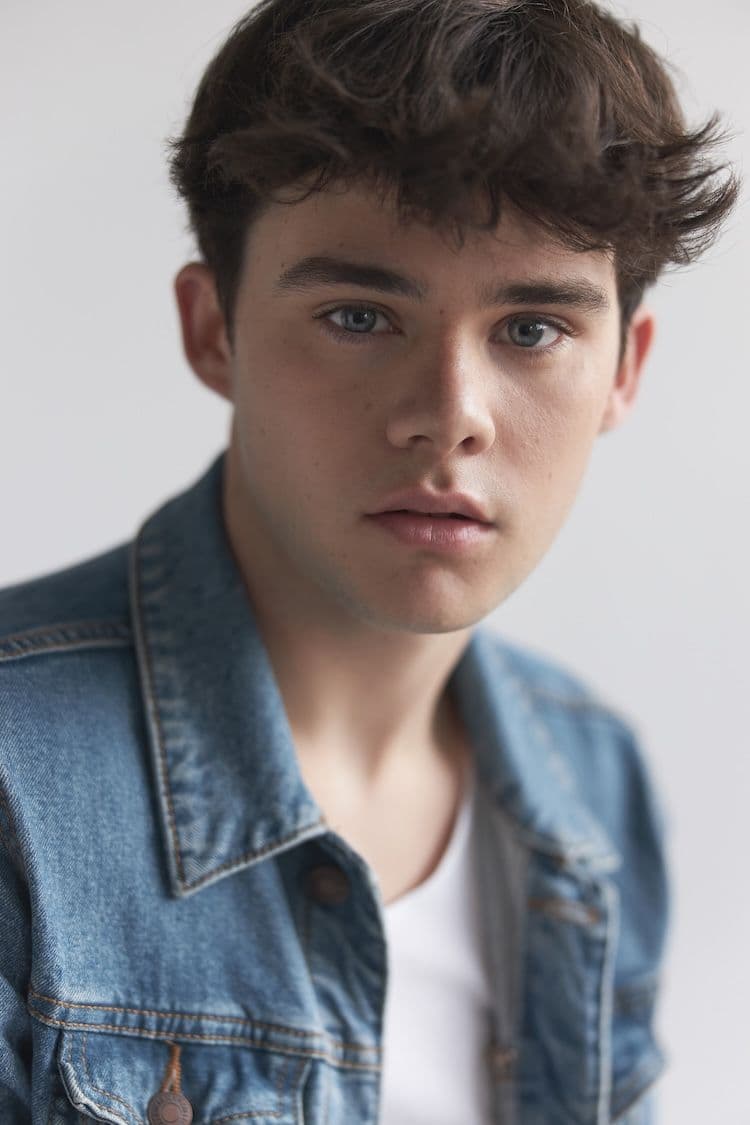 A close-up portrait of actor Ewan Herdman from featuring a young man with messy, textured brown hair and light blue-green eyes looking forward with a neutral expression. He is wearing a blue denim jacket over a white undershirt against a bright, out-of-focus white background.