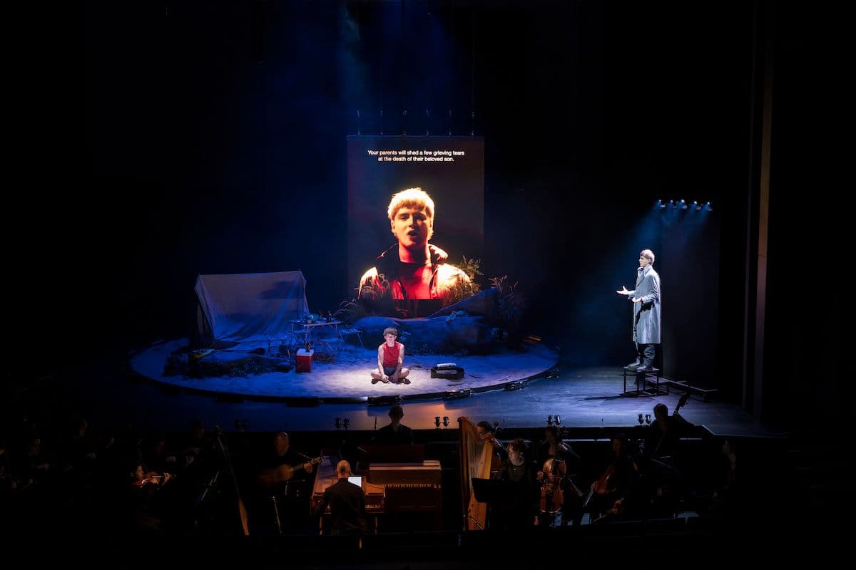 Actor Ewan Herdman as cain dressed in a red cut-off t-shirt sits in a circular pit filled with sand. Baritone Freddy Shaw stands on a stool side stage, with his image projected larger than life, behind Ty, as Lucifer,