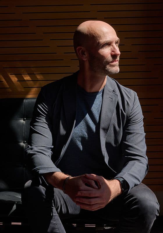 A portrait from the file Erin headshot featuring a bald man with a short beard sitting in a black leather chair. He is captured in profile, looking off to the right into bright sunlight. He wears a grey blazer over a blue t-shirt and dark pants, posed against a slatted wooden wall background.