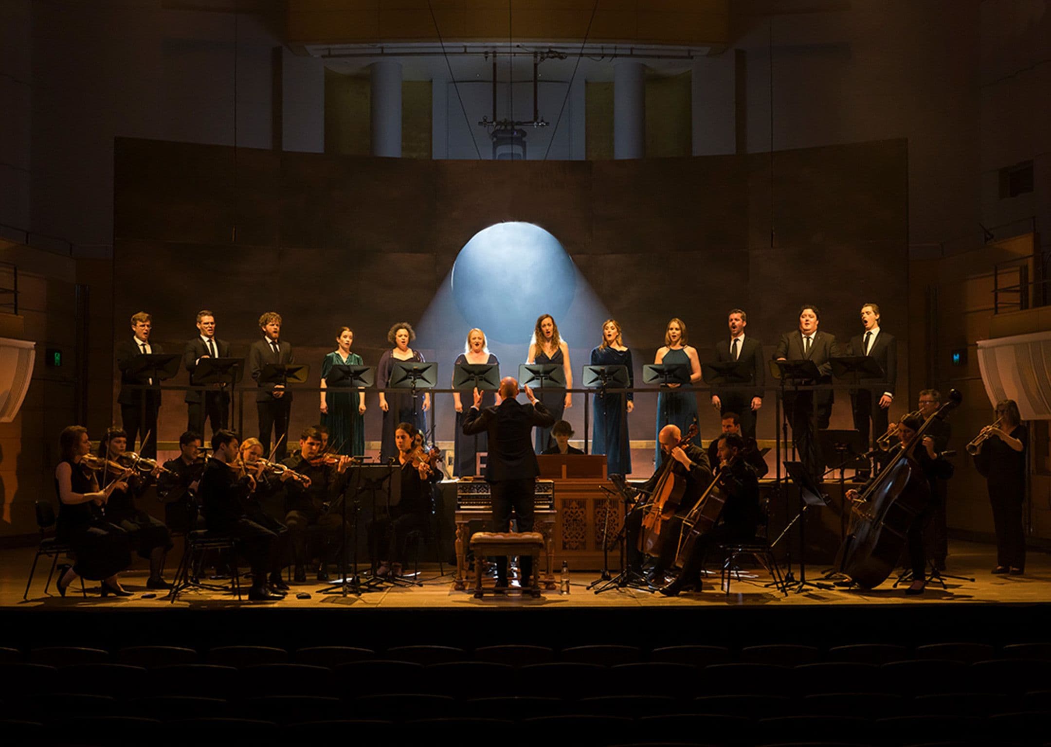 a group of 12 singers stand behind an orchestra with an illuminated circle cutout of the backdrop