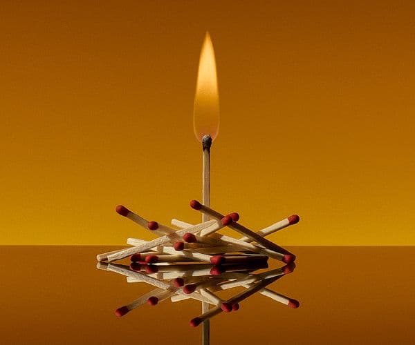 A pile of red-tipped matchsticks against an orange background. One matchstick stands upright in the centre, burning brightly.