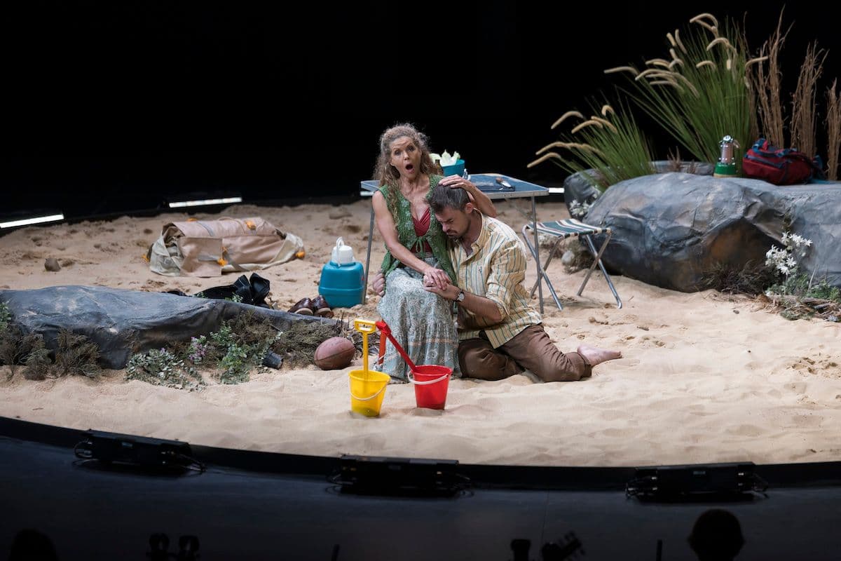 Soprano Sara Macliver as Eve, cradles tenor Kyle Stegall as Adam in Pinchgut Opera's The First Murder. They sit on circular stage filled with sand, representing a beach heath, with a couple of large rocks with grass